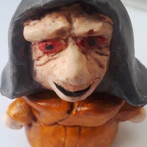 May include: A small ceramic figurine of a person wearing a brown robe and a black hooded cloak. The figurine has a pale face with red eyes and a mischievous smile.