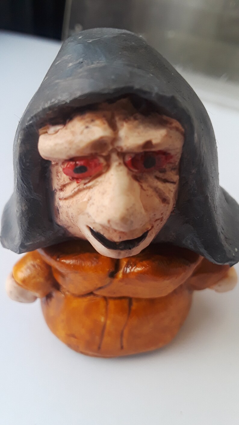 May include: A small ceramic figurine of a person wearing a brown robe and a black hooded cloak. The figurine has a pale face with red eyes and a mischievous smile.