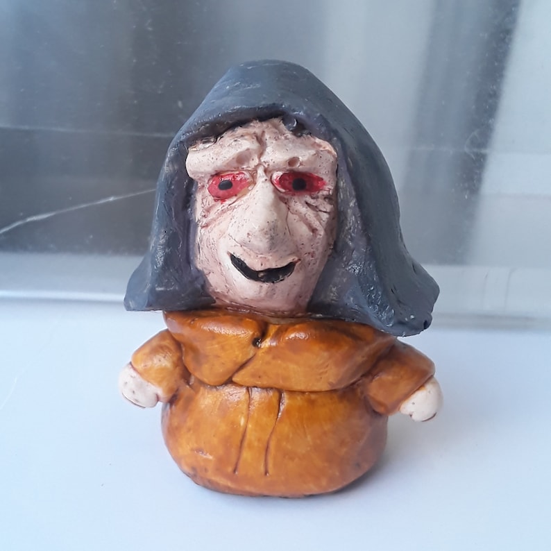 May include: A small ceramic figurine of a person wearing a dark grey hooded robe and a brown tunic. The figurine has a painted face with red eyes and a mischievous smile.