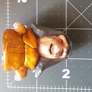 May include: A hand-painted ceramic figurine of a person with a brown jacket, gray hair, and red eyes. The figurine is wearing a brown jacket and has a detailed facial expression.