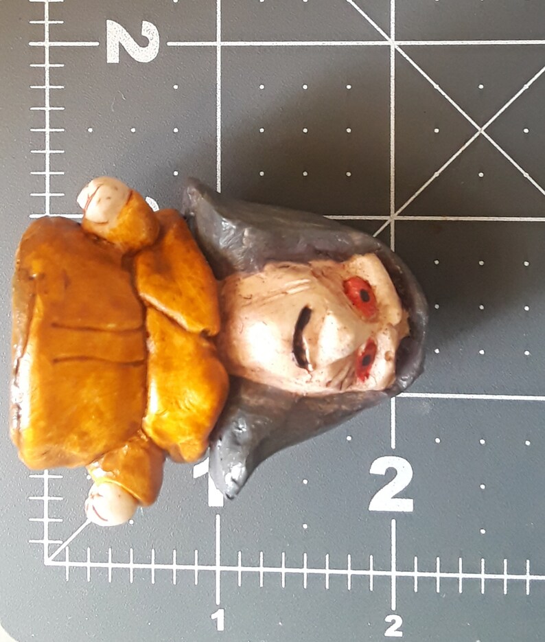 May include: A hand-painted ceramic figurine of a person with a brown jacket, gray hair, and red eyes. The figurine is wearing a brown jacket and has a detailed facial expression.