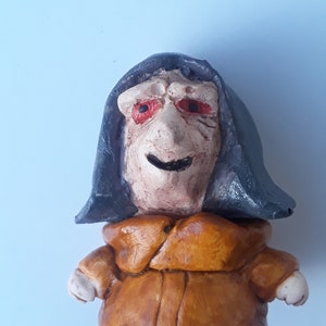May include: A small ceramic figurine of a person wearing a dark blue hooded robe and a brown tunic. The figurine has a smiling face with red eyes.
