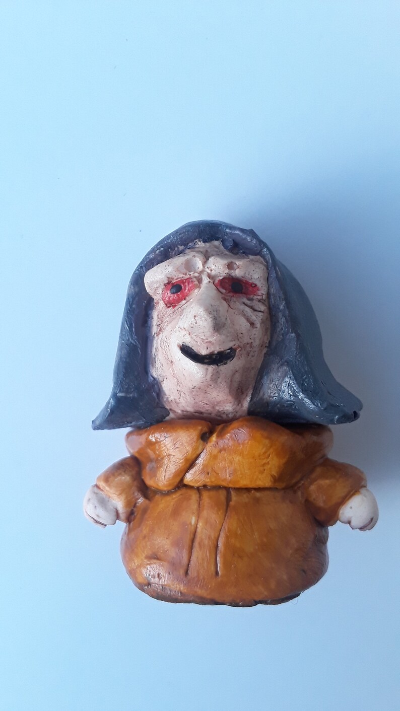 May include: A small ceramic figurine of a person wearing a dark blue hooded robe and a brown tunic. The figurine has a smiling face with red eyes.