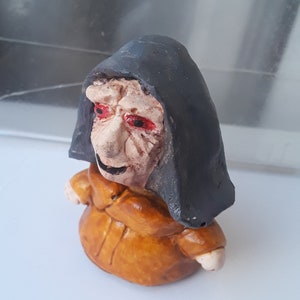 May include: A small ceramic figurine of a witch with a black hooded cloak and a brown robe. The witch has a wrinkled face with red eyes and a mischievous smile.
