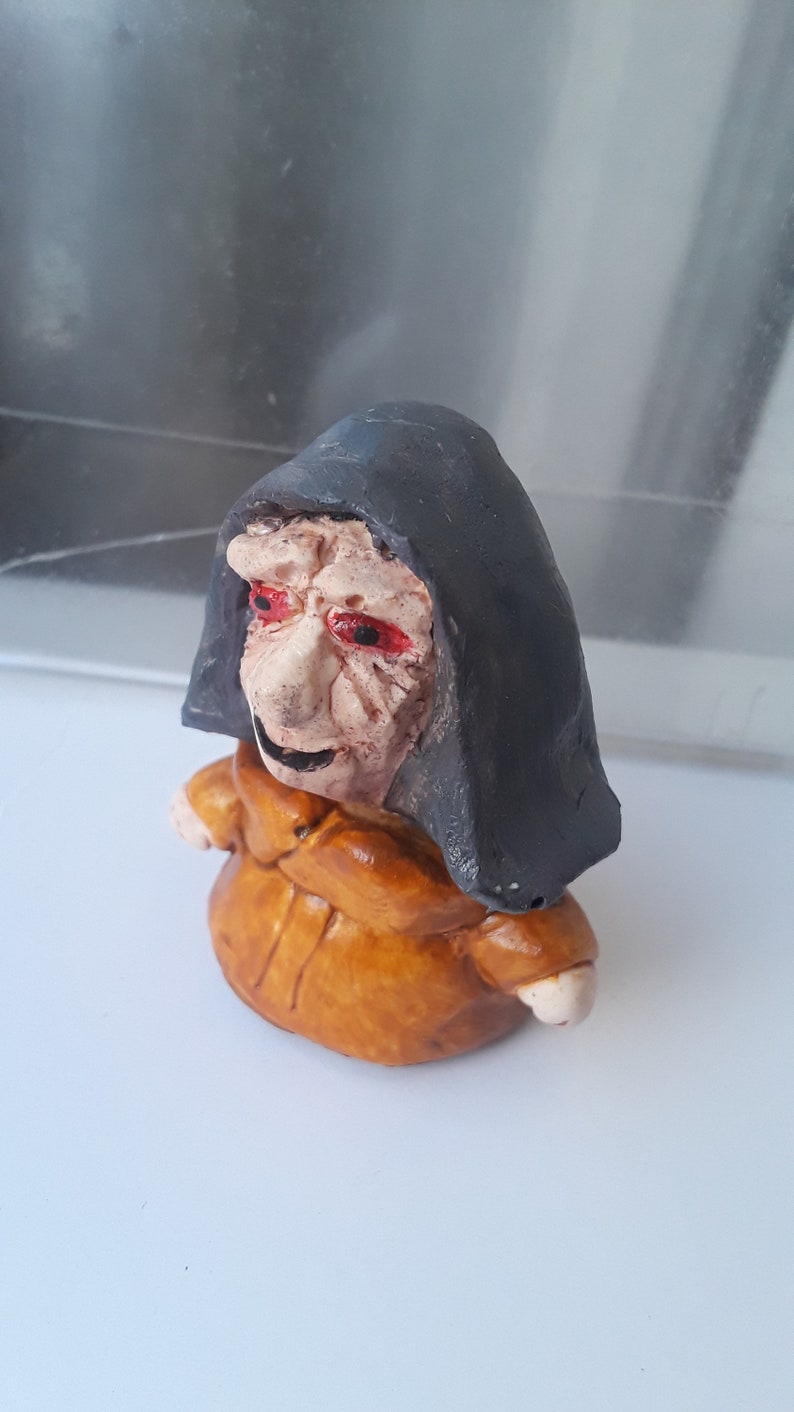 May include: A small ceramic figurine of a witch with a black hooded cloak and a brown robe. The witch has a wrinkled face with red eyes and a mischievous smile.