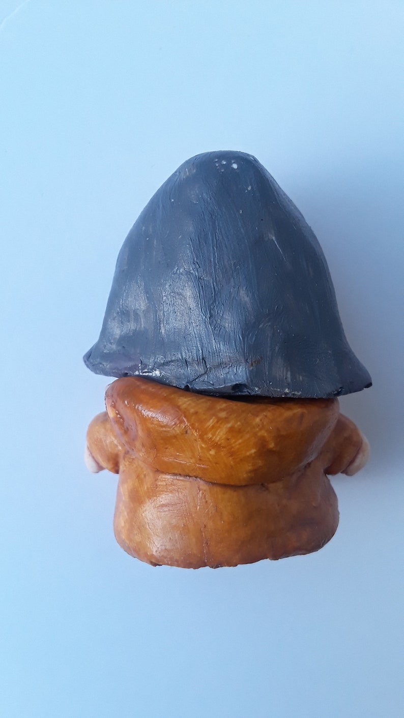 May include: A small clay figurine of a person wearing a brown hooded jacket and a gray hat.