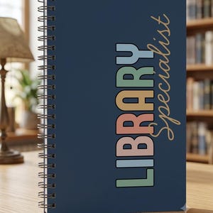 May include: A dark blue spiral-bound notebook featuring the word LIBRARY in stacked, multi-coloured block letters including light blue, sage green, and soft pink, with the word Specialist written in a gold cursive script to the right side.