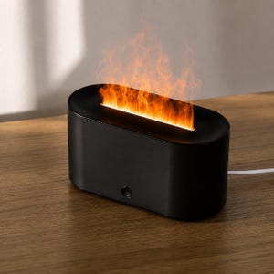 May include: A black, oval electric fireplace with a simulated flame effect. The device is on a wooden surface and has a single button on the front. A white power cable extends from the back.