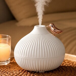 Pu&ograve; includere: Un diffusore per aromaterapia bianco con un design a coste e un accento di uccello in oro rosa. Il diffusore emette un flusso di vapore. Una candela accesa in un barattolo di vetro &egrave; in primo piano.