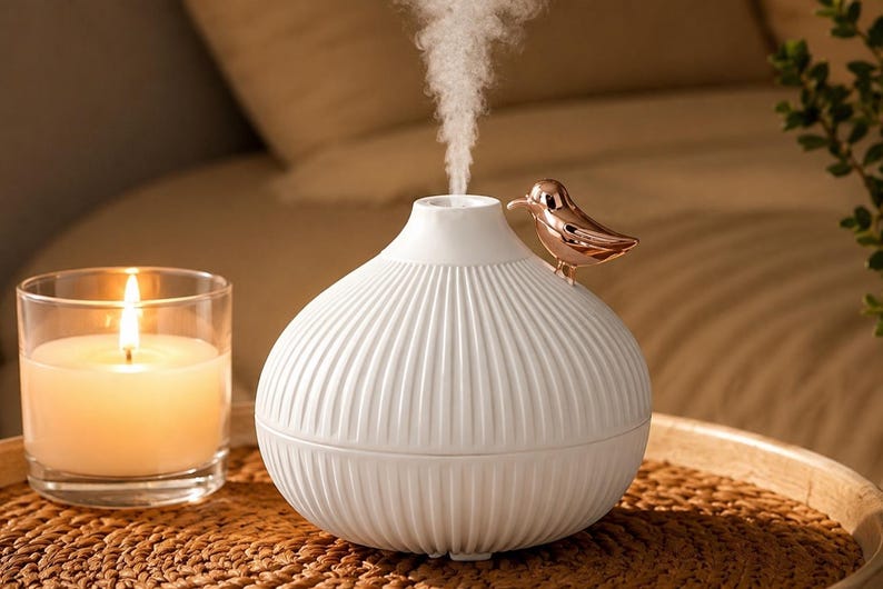 Pu&ograve; includere: Un diffusore per aromaterapia bianco con un design a coste e un accento di uccello in oro rosa. Il diffusore emette un flusso di vapore. Una candela accesa in un barattolo di vetro &egrave; in primo piano.
