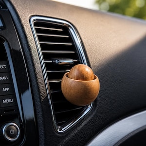 Può includere: Un profumatore per auto in legno con una sfera profumata rotonda in legno naturale all'interno di una piccola ciotola di legno. Il profumatore è posizionato in una bocchetta dell'auto, con un cruscotto nero e finiture argentate sullo sfondo.