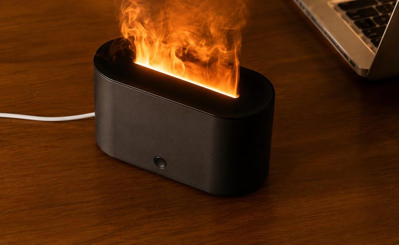 May include: A black, rectangular electric fireplace with a flame effect. The device has a rounded top and a single button on the front. A white power cord extends from the back. The flame effect emits orange and yellow light.