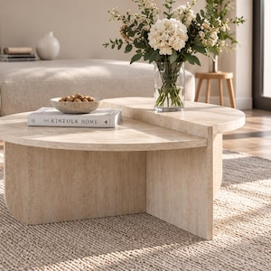 Può includere: Un tavolino da caffè in travertino beige con un design circolare a due livelli e una base spessa e curva. Sul tavolo si trovano un libro bianco intitolato The Kinfolk Home, una piccola ciotola piena di noci e un vaso di vetro trasparente.