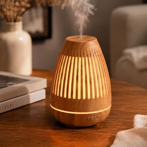 Op de afbeelding: Een houten diffuser voor etherische olie met een lichtbruine afwerking. De diffuser heeft verticale sleuven die een warme gloed uitstralen. Stoom stijgt op van de bovenkant, wat aangeeft dat hij in gebruik is. Hij staat op een ronde houten tafel, wat duidt op een woondecoratie-item.