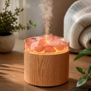 Peut inclure: Un diffuseur d'huiles essentielles en bois avec un dessus en verre rempli de cristaux de sel rose. Le diffuseur émet un jet de vapeur. La base porte le texte "H2O" et un motif de grain de bois. Le diffuseur est sur une surface en bois.