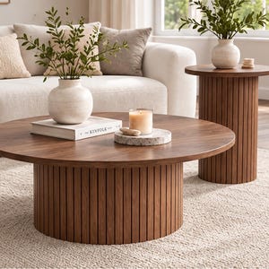 Pode incluir: Uma mesa de centro redonda e uma mesa lateral a condizer, ambas com acabamento em madeira castanha escura e base cilíndrica com ripas verticais. A mesa de centro contém um vaso branco com vegetação, um livro chamado The Kinfolk e uma vela.