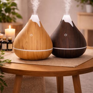Puede incluir: Dos difusores de aceites esenciales, uno de madera clara y otro marrón oscuro, sobre una mesa redonda de madera. Ambos tienen una parte superior blanca donde se libera vapor. Una línea blanca rodea la base de cada difusor. El fondo incluye plantas y una vela encendida.