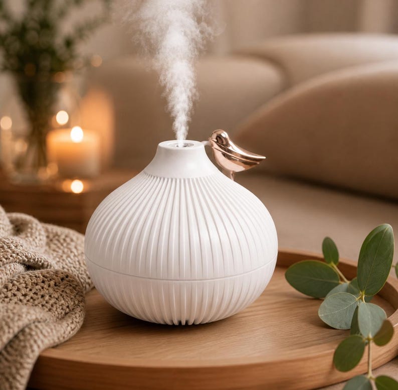 Pu&ograve; includere: Un diffusore di oli essenziali bianco con un design a coste e un accento di uccello dorato. Il diffusore emette un flusso di vapore. Si trova su un vassoio di legno con foglie di eucalipto, creando un'atmosfera da spa.