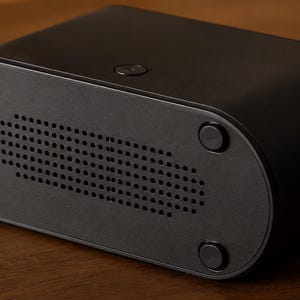 May include: A black electronic device with a rectangular body and rounded ends. The base has a grid of small holes and four circular feet. A small button is on the top. The device is likely a media player or similar, measuring approximately 15 cm in length.