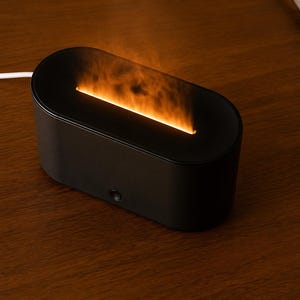 May include: A black, oval aromatherapy diffuser with an orange flame effect. The diffuser is on a wooden surface. A white power cord extends to the left. The device is approximately 20 cm long.