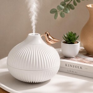 Pu&ograve; includere: Un diffusore di oli essenziali bianco con un design a coste e un accento a forma di uccello dorato. Il diffusore emette un flusso di vapore. Una piccola pianta succulenta in vaso e un vaso decorativo sono sullo sfondo.