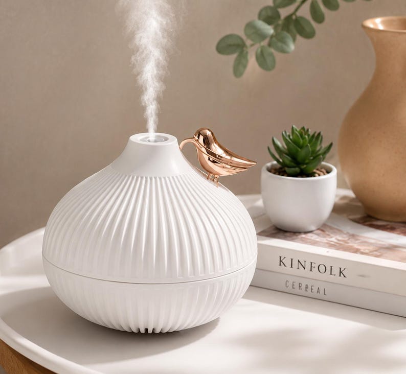 Pu&ograve; includere: Un diffusore di oli essenziali bianco con un design a coste e un accento a forma di uccello dorato. Il diffusore emette un flusso di vapore. Una piccola pianta succulenta in vaso e un vaso decorativo sono sullo sfondo.