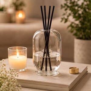 K&ouml;nnte beinhalten: Ein klarer Glasdiffusor mit schwarzen St&auml;bchen, gef&uuml;llt mit Fl&uuml;ssigkeit, steht auf einem neutralfarbenen Buch. Eine brennende Kerze in einem Glashalter und ein kleiner goldener Kerzenhalter befinden sich in der N&auml;he.