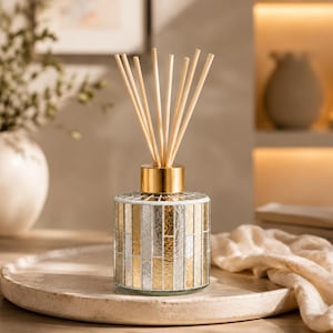Op de afbeelding: Een decoratieve rietverspreider met een goudkleurige metalen kraag en een glazen fles in mozaïekstijl in goud- en zilverkleuren. De diffuser staat op een ronde, lichtgekleurde tray, met houten rietjes die omhoog steken.