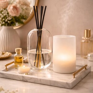 K&ouml;nnte beinhalten: Eine ruhige Anordnung mit einem transparenten Glasdiffusor mit schwarzen St&auml;bchen, einer brennenden Kerze und einer kleinen Kerze auf einem Marmortablett. Eine Vase mit rosa Rosen und Parf&uuml;mflaschen vervollst&auml;ndigen die Szene.
