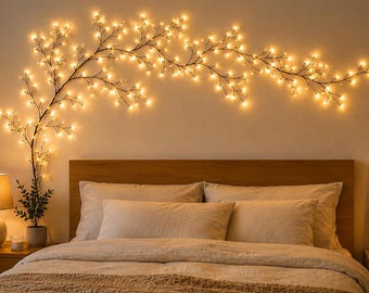 May include: A bedroom with a bed, white bedding, and a wooden headboard. A decorative branch of warm white fairy lights is on the wall. A bedside table with a lamp and a framed print are also visible.