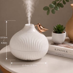Pu&ograve; includere: Diffusore di oli essenziali in ceramica bianca con un ornamento a forma di uccello dorato. Il diffusore &egrave; alto 12,5 cm e largo 13,5 cm. Ha un design a coste ed emette una sottile nebbia. Una piccola pianta succulenta e un libro sono sullo sfondo.