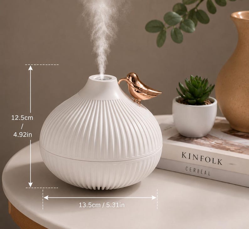 Pu&ograve; includere: Diffusore di oli essenziali in ceramica bianca con un ornamento a forma di uccello dorato. Il diffusore &egrave; alto 12,5 cm e largo 13,5 cm. Ha un design a coste ed emette una sottile nebbia. Una piccola pianta succulenta e un libro sono sullo sfondo.