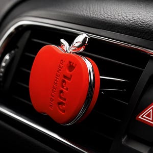 Peut inclure: Diffuseur de parfum pour voiture en forme de pomme rouge, avec des détails argentés. Le mot "APPLE" est gravé sur le devant. L'intérieur de la voiture est noir. Le diffuseur est fixé à une grille d'aération.