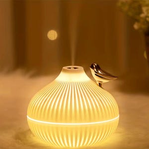 Pu&ograve; includere: Diffusore di oli essenziali color crema con un design a coste e un uccello color oro appollaiato sopra. Il diffusore emette una sottile nebbia e la base si illumina con una luce soffusa. Il design &egrave; elegante e moderno.