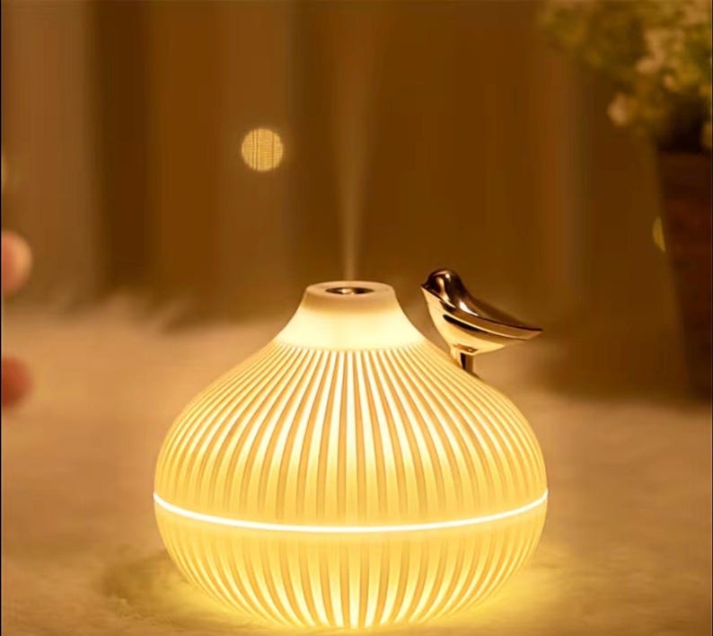 Pu&ograve; includere: Diffusore di oli essenziali color crema con un design a coste e un uccello color oro appollaiato sopra. Il diffusore emette una sottile nebbia e la base si illumina con una luce soffusa. Il design &egrave; elegante e moderno.