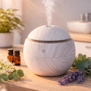 Op de afbeelding: Een witte en grijze diffuser voor etherische olie met houtnerf en stoom die opstijgt. Twee kleine bruine flesjes en takjes lavendel en groen liggen op een houten oppervlak. De diffuser heeft een aan/uit-knop.