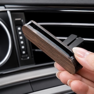 Peut inclure: Un diffuseur de parfum pour voiture avec une finition en grain de bois foncé. Le diffuseur rectangulaire est doté d'un clip noir pour se fixer à une grille d'aération de voiture. L'arrière-plan montre le tableau de bord et les bouches d'aération d'une voiture.