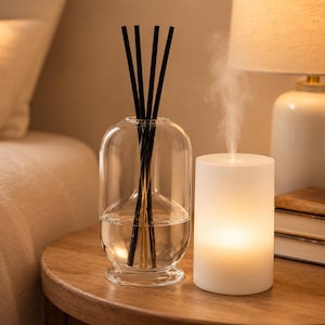 K&ouml;nnte beinhalten: Eine transparente Glasdiffusorflasche mit schwarzen St&auml;bchen und Fl&uuml;ssigkeit, neben einem wei&szlig;en, beleuchteten, zylindrischen Aromatherapie-Diffusor. Die Artikel stehen auf einem Holztisch, mit einer Lampe und B&uuml;chern im Hintergrund. Die Szene wirkt entspannend.