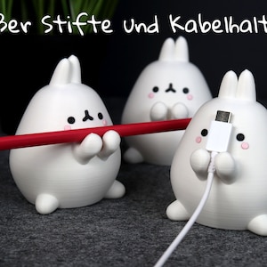 Op de afbeelding: Drie witte bureau-organizers in de vorm van konijnen. Eén houdt een rode potlood vast, een andere een witte USB-C-kabel, en de derde is leeg. De organizers hebben zwarte ogen, roze wangen en kleine armen. De tekst bovenaan luidt "Süßer Stifte und Kabelhalter."