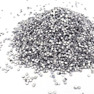 Könnte beinhalten: Ein großer Haufen kleiner, silberfarbener, flach gerückter, runder Verzierungen, die auf einer weißen Oberfläche verstreut sind. Die Verzierungen sind reflektierend und scheinen aus Metall zu sein. Wahrscheinlich für Bastelarbeiten oder Dekoration verwendet.