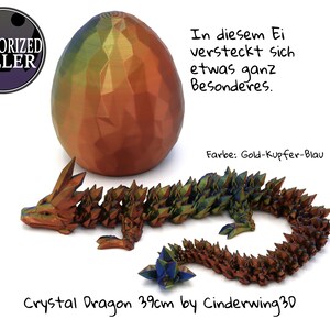 Könnte beinhalten: Eine 39 cm lange, irisierende, goldene, kupferne und blaue Kristalldrachenfigur. Der Drache ist in einer Ruheposition zusammengerollt. Der Drache liegt neben einem großen, irisierenden, goldenen, kupfernen und blauen Ei. Der Text "In diesem Ei versteckt sich etwas ganz Besonderes. Farbe: Gold-Kupfer-Blau" ist in Deutsch auf weißem Hintergrund geschrieben. Der Text "Crystal Dragon 39cm by Cinderwing 3D" ist in Schwarz auf weißem Hintergrund geschrieben.
