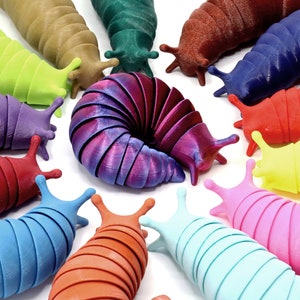 Könnte beinhalten: Eine Sammlung von 12 bunten, segmentierten Schnecken, jede mit einer anderen Farbe und Textur. Die Schnecken sind in einem Kreis um eine größere, schillernde lila Schnecke in der Mitte angeordnet.