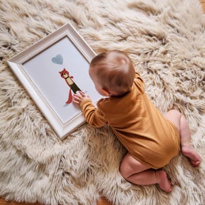May include: A baby in a brown onesie is looking at a framed print of a fox holding a blue balloon. The print is on a white fluffy surface.