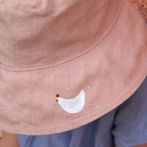 Könnte beinhalten: Ein rosafarbener Eimerhut aus Leinen mit einem weißen gestickten Hühnermotiv.