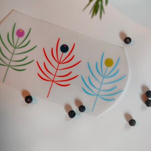 Pode incluir: Uma bandeja retangular branca com três desenhos florais estilizados. Os desenhos são em verde, vermelho e azul com acentos pretos e amarelos. A bandeja está sobre uma superfície branca.