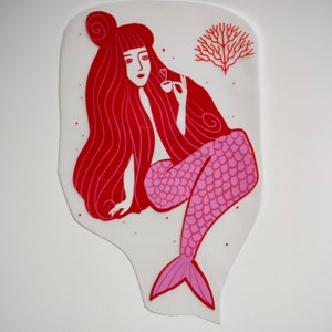 Op de afbeelding: Een rode en roze zeemeermin met lang rood haar houdt een roze kopje vast met een hart erop. De zeemeermin zit op een witte achtergrond met een rode koraaltak achter haar.