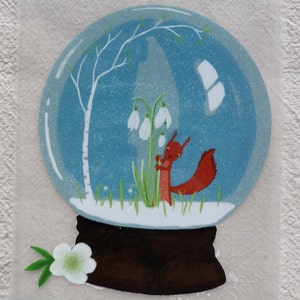 Könnte beinhalten: Eine Schneekugel mit blauem Hintergrund, einem weißen Birkenbaum, einem roten Eichhörnchen und weißen Schneeglöckchen. Der Sockel der Schneekugel ist braun.