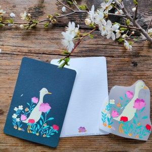 Può includere: Tre carte con un'illustrazione di anatra bianca su sfondo blu, una carta bianca e una carta trasparente con un'illustrazione di anatra bianca su sfondo rosa. Le carte hanno un design floreale con fiori rosa, blu e gialli.