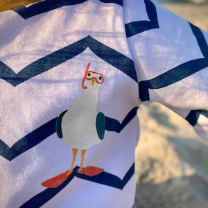 Puede incluir: Camiseta blanca de manga larga con un patrón de chevron azul y blanco. Un gaviota con gafas rojas está impreso en la parte delantera de la camiseta. El texto "Koten&Konsorten" está impreso en la manga derecha.