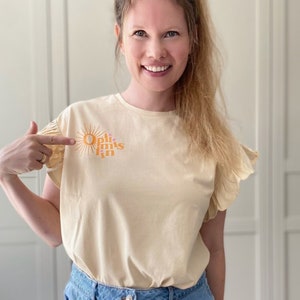 Könnte beinhalten: Eine Frau trägt ein hellbeiges T-Shirt mit einem Grafikdruck einer Sonne und dem Text "Optimismus" in Rosa und Orange. Sie zeigt auf den Druck auf ihrem Shirt.